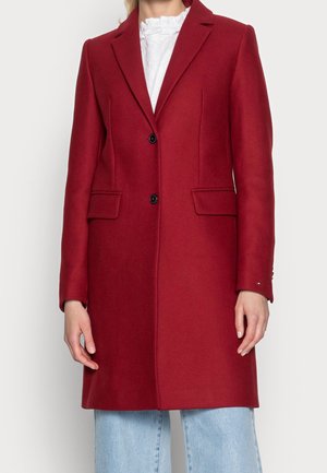 Kobieta ubrana w dopasowany, czerwony płaszcz do kolan z dwoma czarnymi guzikami i z klapkami, na białej bluzce z falbankami i jasnoniebieskich dżinsach.