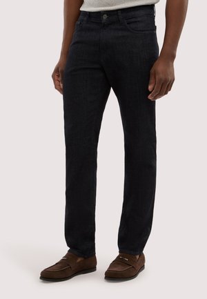 Mand iført mørkeblå jeans med lige ben og brune loafers i ruskind, stående mod en ensfarvet lys baggrund.