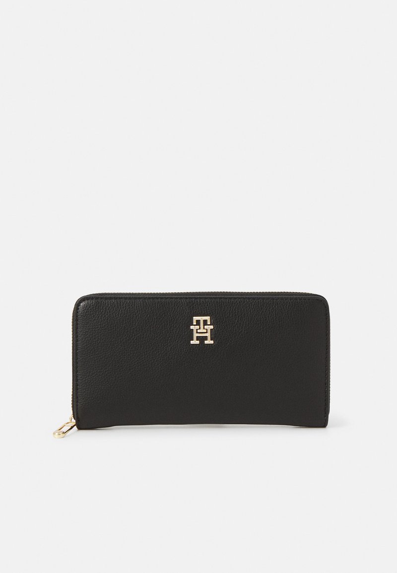 Carteira de couro preto com uma superfície texturizada, apresentando um emblema em ouro e um fecho zipper ao longo da borda superior.