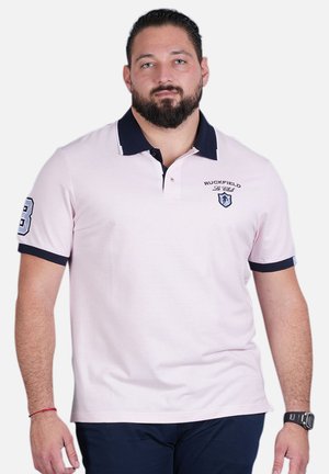 Polo shirt rose clair avec un col bleu marine, à manches courtes et un logo brodé sur la poche gauche. Comprend le numéro 83 sur la manche.