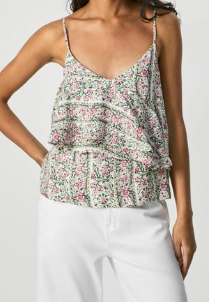 Kobieta w bezrękawniku w kwiatowy wzór z falbankami i cienkimi ramiączkami, w połączeniu z białymi spodniami, na jednolitym tle.