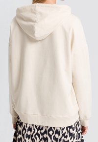 Beige oversized hoodie with a drawstring hood, ribbed cuffs, and hem, made from soft cotton material; worn over a patterned skirt.