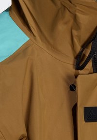 Veste à capuche marron avec bouton noir et écusson logo Adidas noir sur la poitrine, sur fond bleu clair.