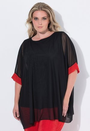 Blonde vrouw draagt een losse zwarte doorschijnende top met rode manchetten en een rode broek, staand tegen een witte achtergrond.
