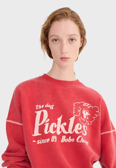 Jeune personne aux cheveux clairs portant un sweat-shirt rouge avec le texte "The dog Pickles - since 09 Bobo Choses" et un dessin animé de chien sur le visage.