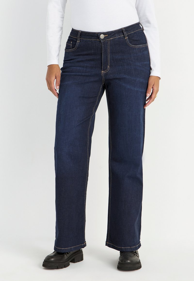 Donkerblauwe denim jeans met een rechte pijp, zichtbare stiksels en twee voorzakken. Gepaard met zwarte schoenen met een dikke zool.