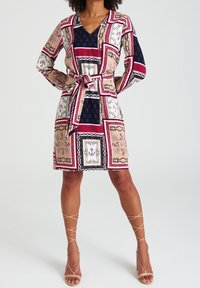 Mehrfarbiges Kleid mit V-Ausschnitt, langen Ärmeln und tailliertem Gürtel. Es zeigt quadratische Muster in Pink, Marineblau und Creme mit dekorativen Akzenten.