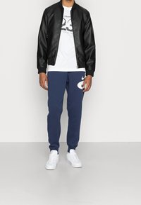 Uomo con giacca in pelle nera, t-shirt grafica bianca, pantaloni sportivi blu navy con logo bianco e sneakers bianche, in piedi su un pavimento grigio.