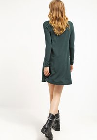 Zielona sukienka z długim rękawem, dopasowanym gorsetem i rozkloszowanym dołem, uszyta z gładkiego materiału. Noszona z czarnymi botkami za kostkę z akcentami łańcuchowymi.