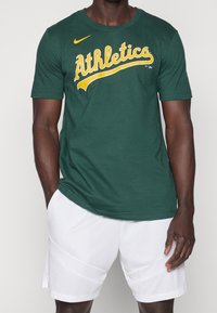 Mørkegrøn bomuld t-shirt med gul "Athletics" tekst, rund hals, korte ærmer, matchende med hvide mesh shorts. Nike-logo på brystet.