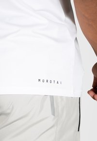 Hvit idretts t-skjorte med en glatt tekstur, har svart "MOROTAI" logo ved kanten, kombinert med lysegrå shorts og detalj med sidelommer.
