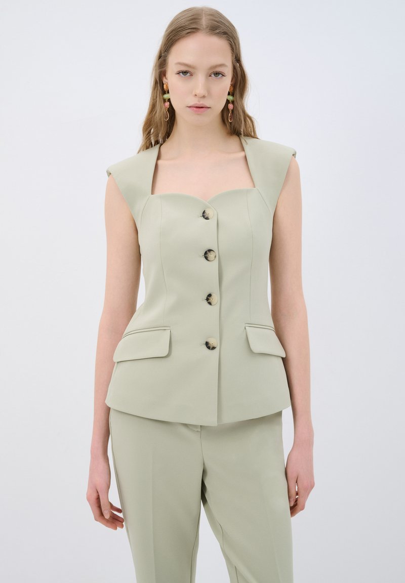 Gilet ajusté vert clair avec de larges bretelles, cinq boutons et poches avant. Texture lisse et silhouette cintrée.