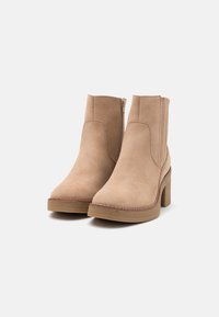 Ett par beige mockaskor i ankelhöjd med sidodragkedjor och blockklackar, stylade mot en vit bakgrund.