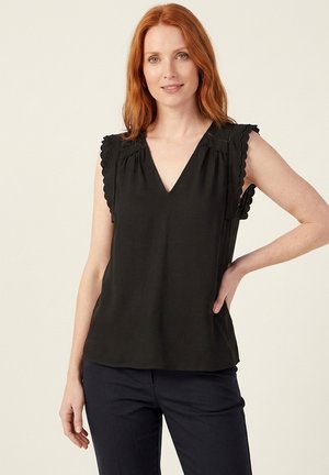 Femme aux cheveux roux portant un haut noir sans manches avec des bords festonnés et un pantalon foncé, debout avec une main sur la hanche sur un fond uni.