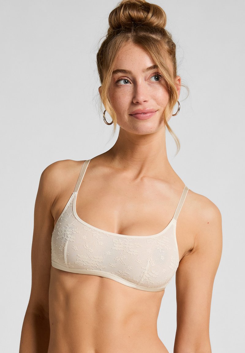 Soutien-gorge en dentelle beige avec des fines bretelles ajustables, une texture douce et des détails de broderie florale sur le tissu. Design simple avec une finition lisse.