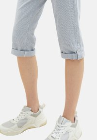 Gambe che indossano pantaloni azzurri arrotolati e scarpe da ginnastica bianche con lacci, dettagli in rete e pelle su uno sfondo bianco.