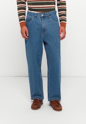 Blue denim jeans with a loose fit, featuring five pockets, a button closure, and stitched accents. Worn with brown leather shoes.
