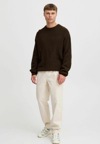 Bruine gebreide trui met een ronde hals, lichtgewicht textuur, gecombineerd met lichtbeige broek en witte sneakers met grijze accenten.