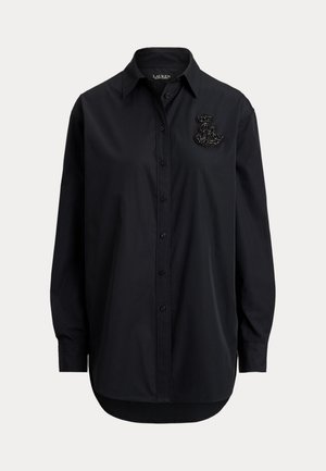 Mujer con una camisa negra de gran tamaño con las mangas remangadas y pantalones blancos de pierna ancha, sosteniendo un pequeño bolso negro con cadena.