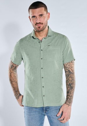 Man with short hair and tattoos on both arms wearing a light green short-sleeve button-up shirt and blue jeans against a plain background.
