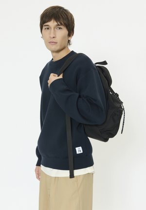 Jonge persoon die een donkerblauw sweatshirt en beige broek draagt, met een zwarte rugzak over één schouder tegen een effen witte achtergrond.