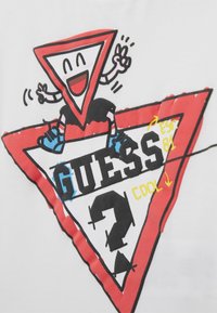 Tegneseriefigur i trekantet form, der smiler og giver et fredstegn, sidder på et større trekantet design med "GUESS" og farverige accenter.