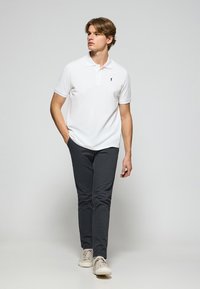 Polo bianco a maniche corte con un piccolo logo nero, abbinato a pantaloni grigio scuro e sneakers chiare, su uno sfondo neutro.