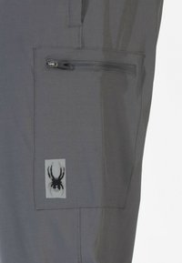 Graue Hose mit einem Reißverschluss-Cargotasche, versehen mit einem schwarzen Spinnenlogo auf einem weißen Hintergrund, aus leichtem Textil gefertigt.