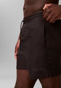 Zwarte zwemboxers met een elastische tailleband, een striksluiting en een reliëf "Calvin Klein" logo aan de zijkant. Gladde, lichte stof.