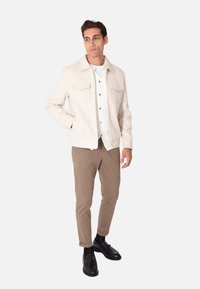 Giacca beige con due tasche sul petto, indossata sopra una camicia bianca con colletto. Pantaloni marroni e scarpe nere con finitura liscia.