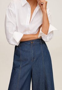 Chemise blanche à manches retroussées, associée à un pantalon en denim bleu foncé à jambes larges, présentant une fermeture à boutons à l'avant et des coutures contrastées.