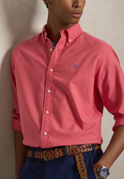 Muž nosící korálovou košili na knoflíky, zapletený hnědý pás, tmavě modré kalhoty a modré hodinky, stojící u obyčejné zdi.