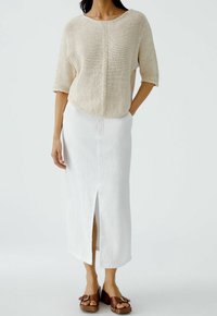 Top lavorato a maglia leggero, beige, con scollo rotondo e maniche corte, abbinato a una lunga gonna bianca in lino con spacco frontale. Sandali con zeppa marroni.