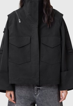 Personne portant une veste noire oversize avec de grandes poches avant et un détail de fermeture éclair diagonal, associée à un jean en denim gris.