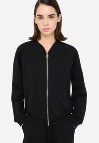 Čierna bomber bunda na zips s raglánovými rukávmi, zlato-zipsom a bočnými vreckami. Mäkka textúra tkaniny so priliehavým spodným lemom.