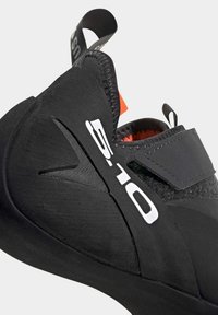 Schwarzer Kletterschuh mit strukturierter Oberfläche, Klettverschluss und weißem Logo. Verfügt über eine Gummisohle und eine Lasche zum einfachen Anziehen.