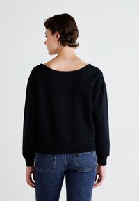 Pessoa com cabelo curto, vestindo um sweatshirt preto sem ombros e jeans azuis, de pé de costas para a câmara contra um fundo simples.