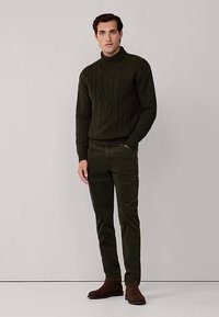 Mörkgrön kabelstickad polotröja, kombinerad med mörkgröna smala byxor och bruna mockaankelstövlar, stående mot en neutral bakgrund.