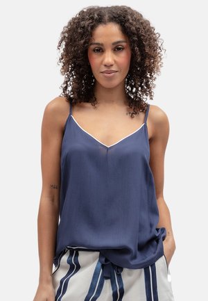 Vrouw met krullend haar die een marineblauwe mouwloze top en witte broek met verticale blauwe strepen draagt, staand tegen een effen achtergrond.