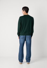 Donkergroen sweatshirt met lange mouwen, geribbelde manchetten en onderrand, gedragen met losse blauwe spijkerbroeken en beige schoenen.