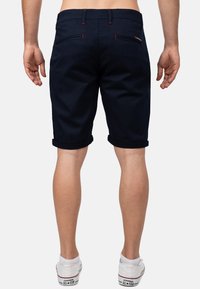 Pantaloni corti blu navy con risvolti, realizzati in tessuto leggero. Presentano dettagli di cucitura rossa e tasche posteriori. Indossati con sneaker bianche.