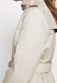 Beżowy płaszcz typu trencz z wysokim kołnierzem, guzikami oraz paskiem w talii. Gładka tkanina, dopasowany krój i wyraźnie zaznaczone ramiona.