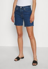 Pantalones cortos de mezclilla en azul oscuro, con una cintura de altura media, cinco bolsillos y un dobladillo vuelto. Combinados con sandalias deslizantes de tejido negro.