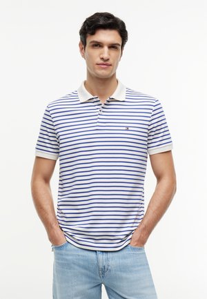 Jeune homme portant un polo blanc avec des rayures horizontales bleues et un jean bleu clair, debout les mains dans les poches devant un fond blanc.
