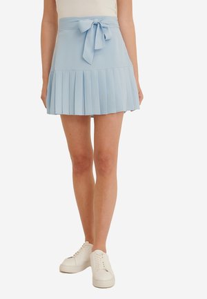 Woman wearing a light blue pleated mini skirt with a tied bow at the waist and white lace-up sneakers, standing against a white background.
