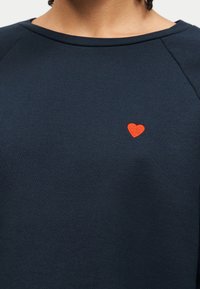 Personne portant une chemise bleu foncé avec un petit cœur rouge brodé sur la poitrine gauche, avec le col et les épaules visibles.