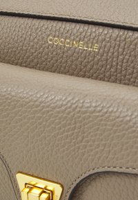 Beige Lederhandtasche mit strukturierten Oberflächen, goldfarbener Hardware-Schließe und geprägtem Branding. Minimalistisches Design mit klaren Linien.