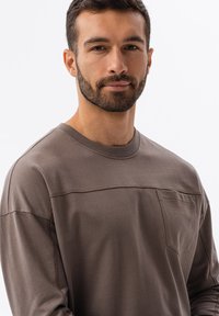 Hnědá košile s dlouhým rukávem vyrobená z měkké tkaniny, s žebrovaným výstřihem, náprsní kapsou a šitým panelem přes přední část.