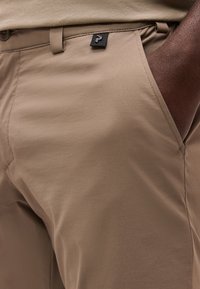 Κοντινό πλάνο του χεριού ενός ατόμου στην μπροστινή τσέπη μπεζ παντελονιού με μια μικρή μαύρη ετικέτα λογότυπου κοντά στη μέση.