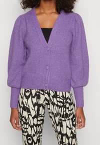 Lila stickad kofta med V-ringning, ribbad textur och puffärmar, ihop med leggings med svart och vitt mönster. Tre knappar framtill.